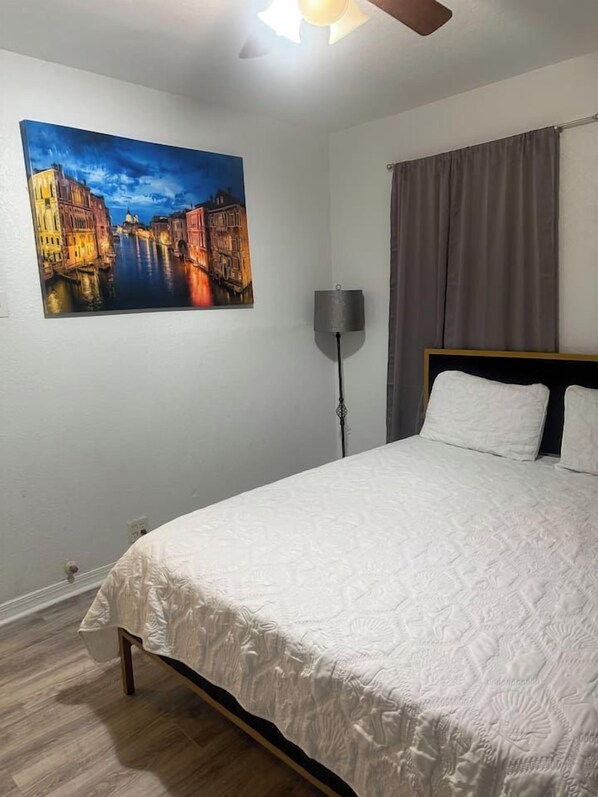 Une chambre à coucher avec un lit, un tableau représentant un paysage urbain, une lampe sur pied et une fenêtre avec des rideaux.