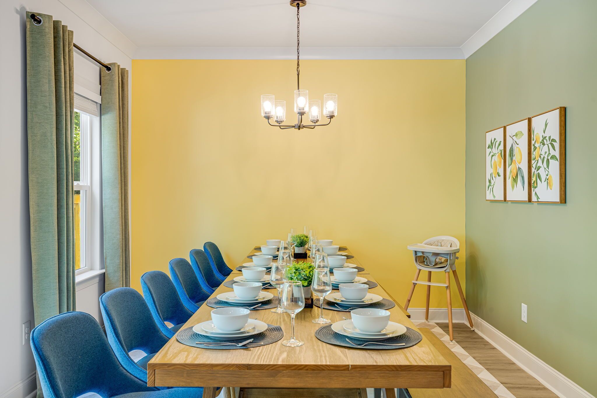 Un comedor con una larga mesa dispuesta para una comida, sillas azules, una araña de cristal y una trona para bebés.