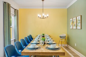 Une salle à manger avec une longue table dressée pour le repas, des chaises bleues, un lustre et une chaise haute pour bébé.