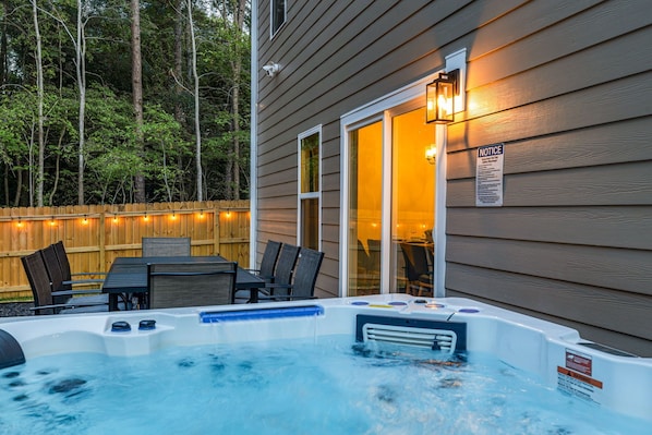 Un bain à remous avec une terrasse en bois et des chaises, une clôture en bois et une maison dont une fenêtre est éclairée.