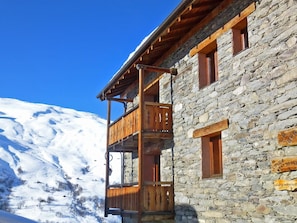 A stone building with a wooden balcony, a clear blue sky, and a snow-covered mountain in the background.
