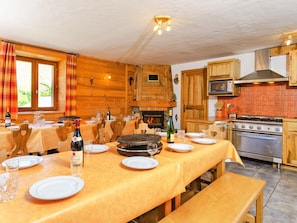 A cozy dining area with a long table set for a meal, a fireplace, wooden cabinets, and a modern kitchen with a stove.