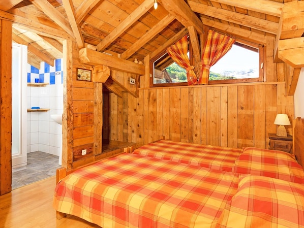 A wooden cabin bedroom with a checkered bedspread, a window with curtains, and a view of the outdoors.