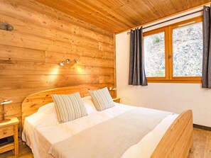 A wooden cabin bedroom with a bed, two bedside tables, a nightstand, a window with curtains, and a wooden ceiling. - Balcon du Paradis by Interhome (Peisey-Nancroix)