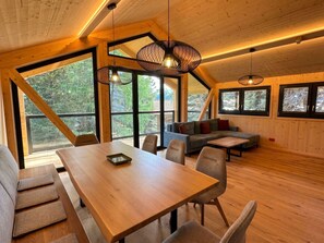 A spacious room with a wooden dining table, chairs, and a sofa. Large windows provide natural light and views of the outdoors. - 02_NTH054_18e_Sup Chalet mit Sauna und Hot Tub 2 E by Interhome (Großveitsch)