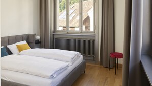 A modern bedroom with a grey upholstered bed, a bedside table with a lamp, a radiator, and a window with curtains. - Auberge Adler, (Fläsch). Doppelzimmer by Interhome (Fläsch)