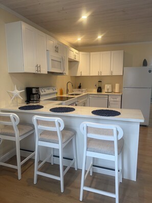 A kitchen with white cabinets, a central island with bar stools, a coffee maker, and a refrigerator. - Coastal Haven Getaway (Treasure Island)