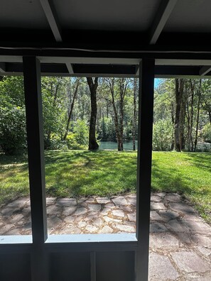 Vista a través de una ventana que muestra un área empedrada y un césped que conduce a una zona boscosa con árboles y una masa de agua.