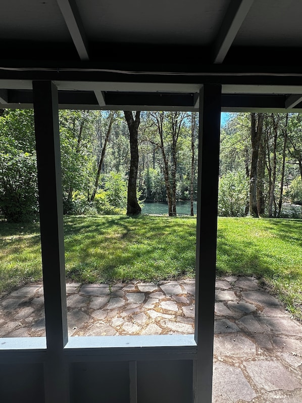 A view through a window with a stone-paved area and a grassy lawn leading to a wooded area with trees and a body of water.