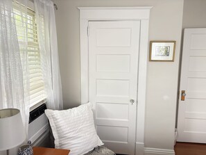 Ein Zimmer mit einer weißen Tür, einem Fenster mit Rollläden, einem weißen Kopfkissen, einem gerahmten Bild an der Wand und einer Heizung.