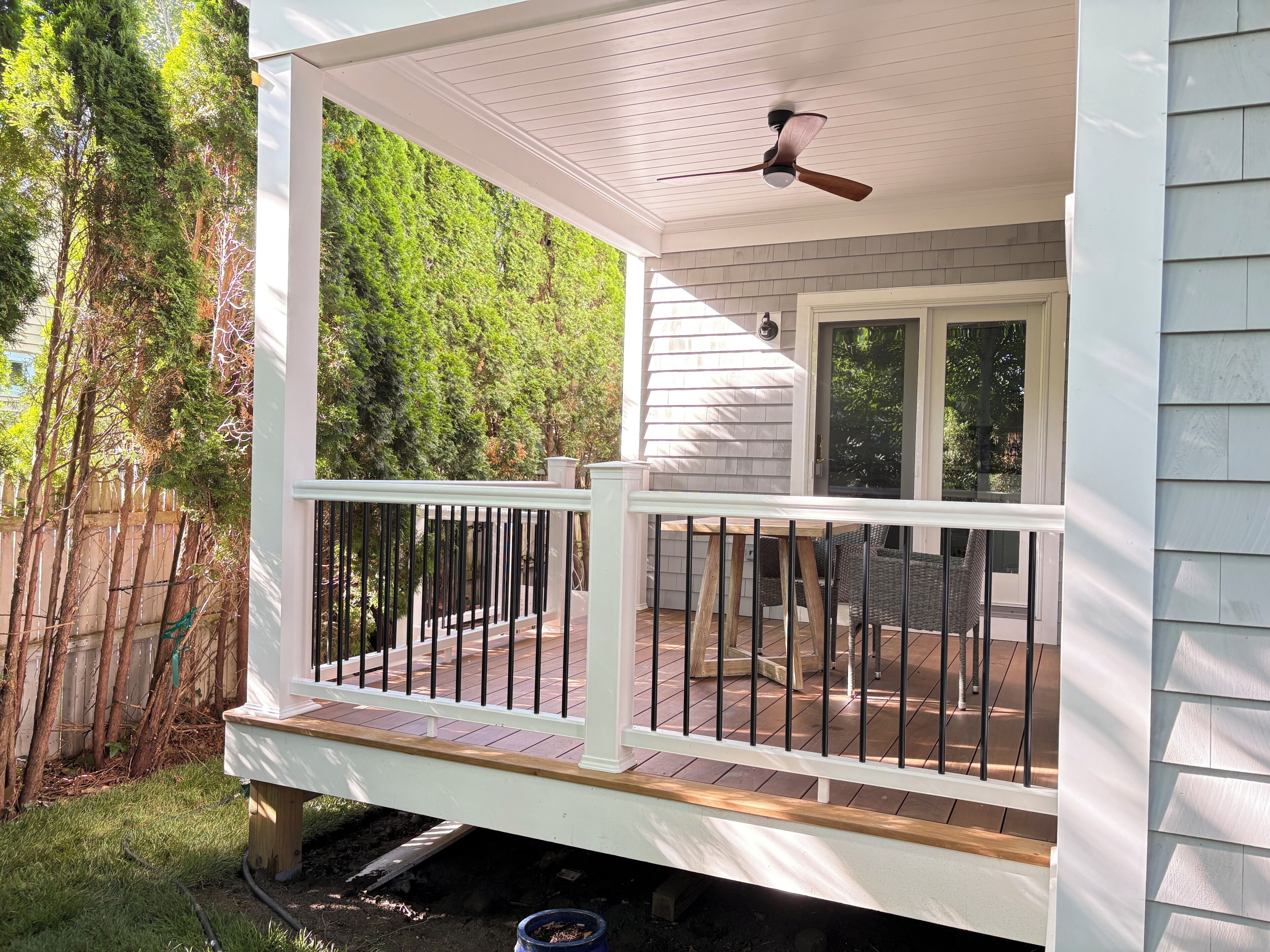 A covered outdoor deck with a railing, a ceiling fan, and a view of a wooded area.