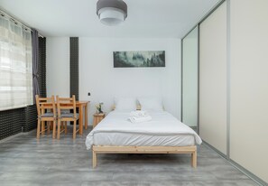 A bedroom with a bed, a desk, chairs, a painting, and a window with curtains.