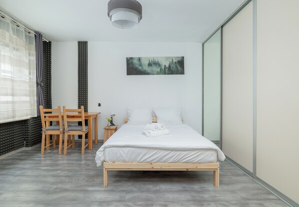 A bedroom with a bed, a desk, chairs, a painting, and a window with curtains.