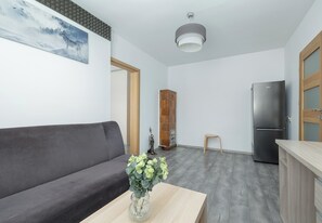A modern living room with a grey sofa, a wooden coffee table with a vase of flowers, a small wooden stool, and a kitchen area with a stainless steel refrigerator.