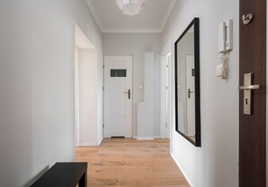 A hallway with a wooden floor, white walls, a door with a window, and a large mirror on the right wall. - Wola Ratusz Comfort Apartment (Warszawa)