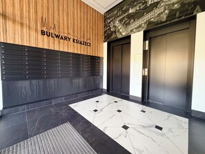 A modern elevator area with a wooden panel wall, a black panel with a number list, and a marble-tiled floor. - Bulwary Książęce Central Apartment (Wrocław)