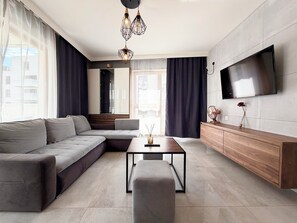 Ein modernes Wohnzimmer mit Eckcouch, Couchtisch, Wand-Fernseher und großem Fenster mit Vorhängen.
