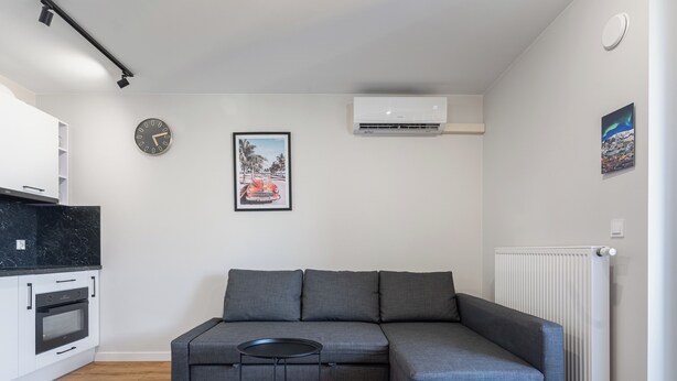 Una sala moderna con un sofá modular, una mesita redonda, un área de cocina con armarios blancos, un reloj, un equipo de aire acondicionado y un radiador.