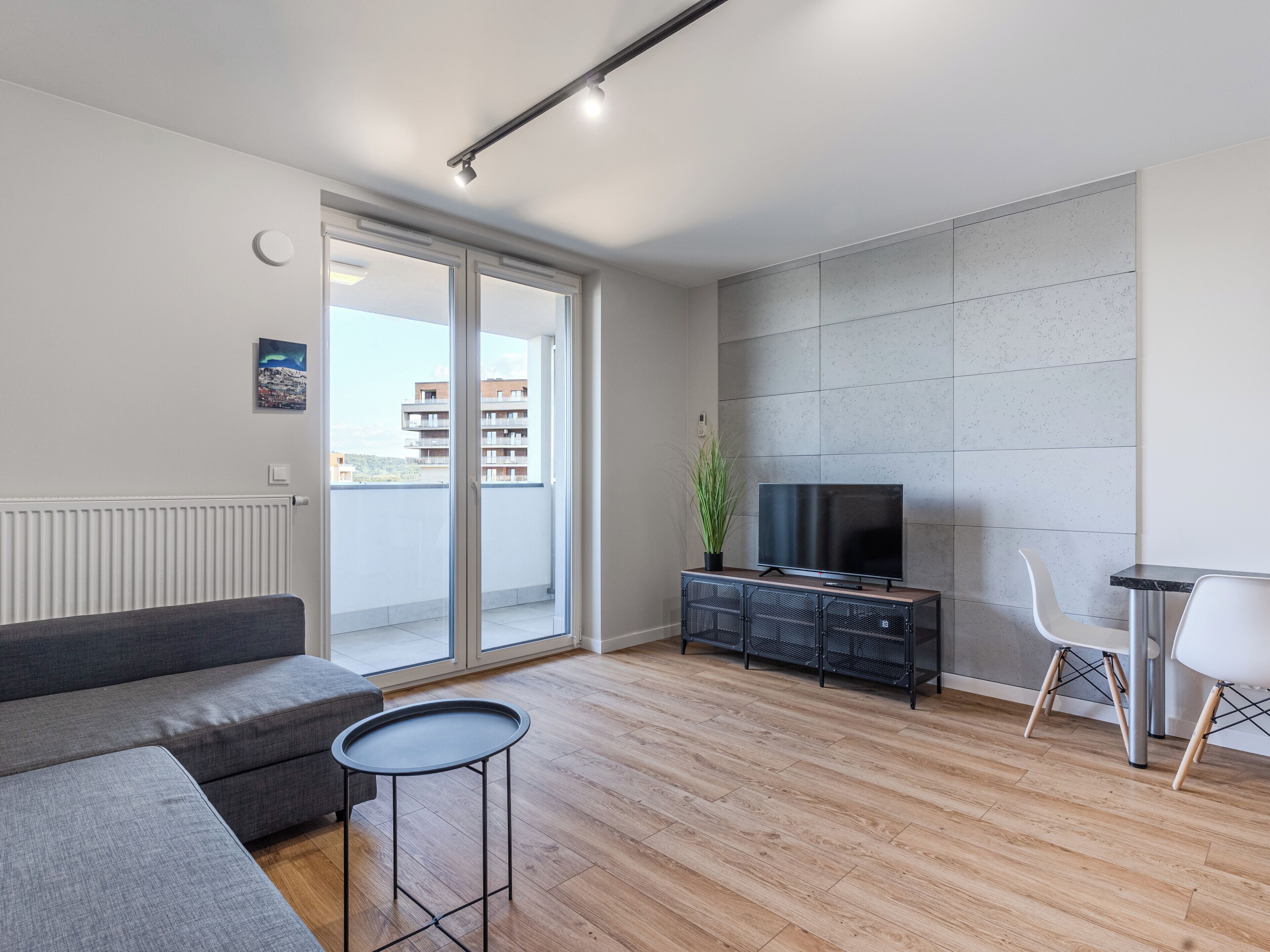 Ein modernes Wohnzimmer mit einem grauen Sofa, einem kleinen runden Tisch, einem Fernseher auf einem schwarzen Schrank und einem Balkon mit Blick auf die Stadt.