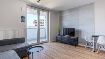 Ein modernes Wohnzimmer mit einem grauen Sofa, einem kleinen runden Tisch, einem Fernseher auf einem schwarzen Schrank und einem Balkon mit Blick auf die Stadt.