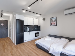 Moderni hotellihuone, jossa on pieni keittiö, valkoisilla lakanoiden peitetty sänky, harmaa pääty ja seinälle asennettu ilmastointilaite.
