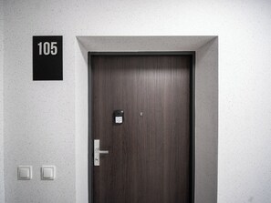 En mörk trädörr med ett digitalt lås och ett nyckelhål, ett nummer ’105’ ovanför, och två vita strömbrytare på väggen.