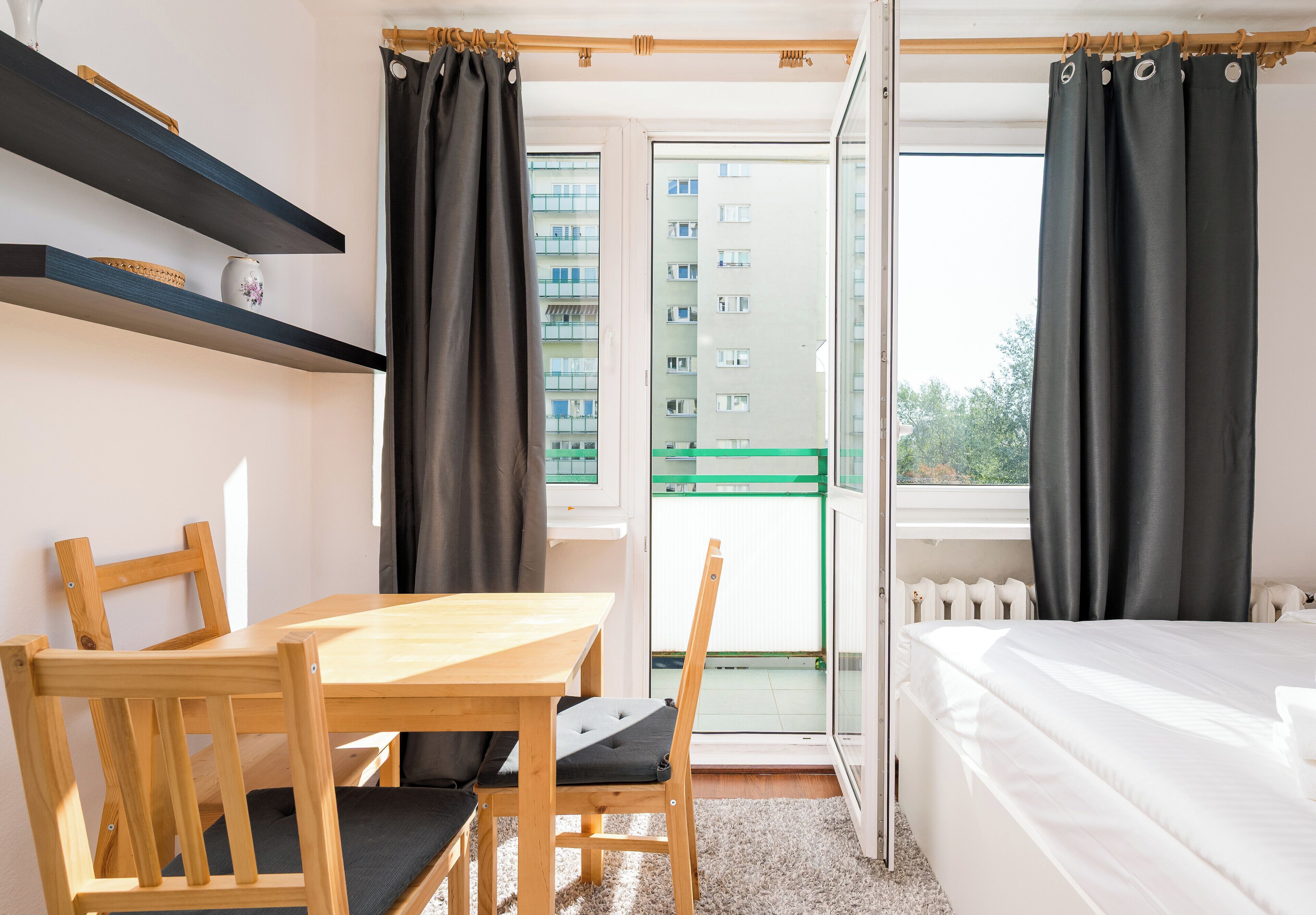 A small, well-lit room with a wooden table, a chair, a bed, and a window with curtains.
