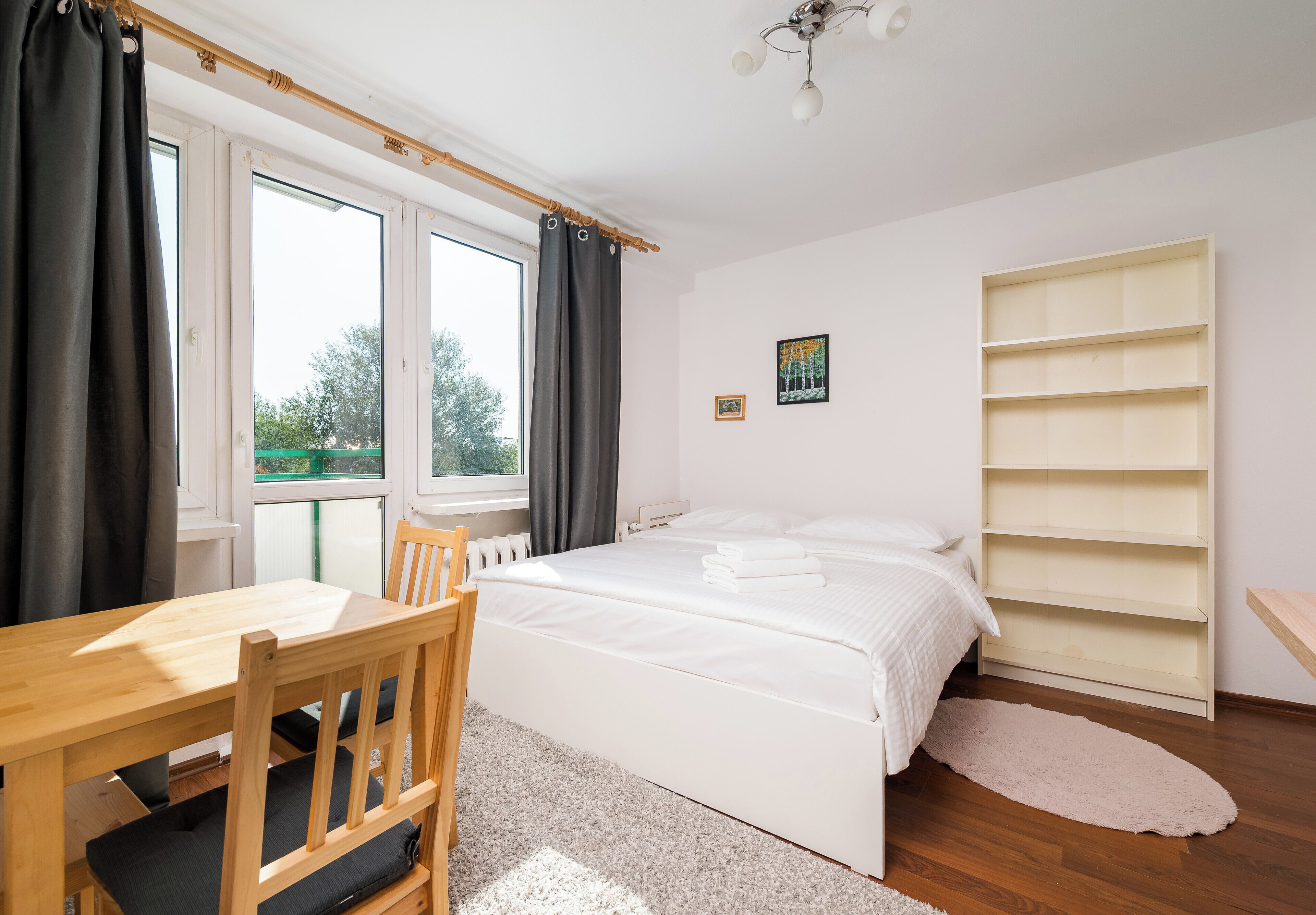 Une chambre à coucher comprenant un lit, une chaise en bois, une table, une fenêtre avec des rideaux, un radiateur, un tableau accroché au mur et une étagère.