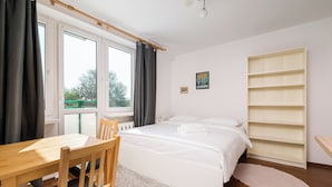 Una camera da letto con un letto, una sedia di legno, un tavolo, una finestra con tende, un termosifone, un quadro appeso al muro e una scaffalatura.