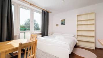 Une chambre à coucher comprenant un lit, une chaise en bois, une table, une fenêtre avec des rideaux, un radiateur, un tableau accroché au mur et une étagère.