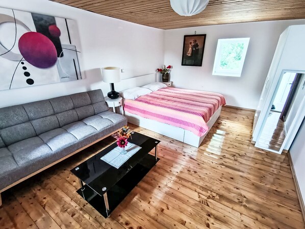Quarto moderno com um sofá cinzento, uma cama com colcha listrada em rosa e branco, uma pequena mesa de cabeceira com uma luminária e um quadro na parede.