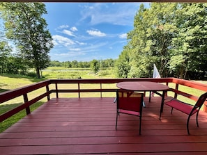 A wooden deck with a table and chairs, overlooking a grassy field and trees.