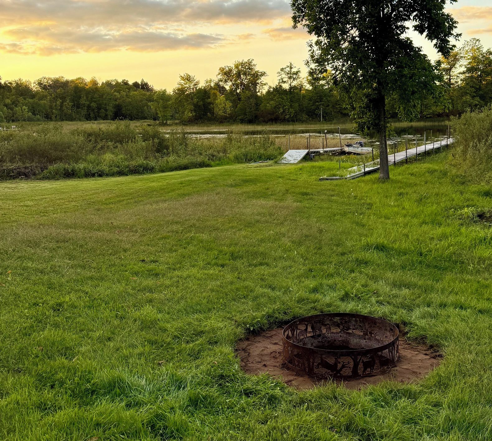 A grassy field with a metal fire pit in the foreground and a dock extending into a body of water in the background.
