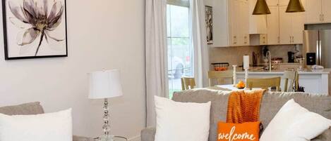 A modern living room with a grey sofa, a coffee table, and a lamp. There is a kitchen in the background with white cabinets and a refrigerator.