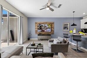 A modern living room with a flat-screen TV, a ceiling fan, and a dining area with a bar. - Brand New | 4Bd Private Pool, Resort Near Disney  4993 (Kissimmee)