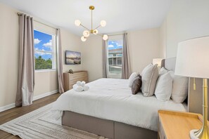 A bedroom with a bed, bedside tables, a lamp, curtains, a TV, and a view of the outdoors. - Brand New 5BR 5BA Villa w Pool, Spa & Game Room 7800 (Kissimmee)