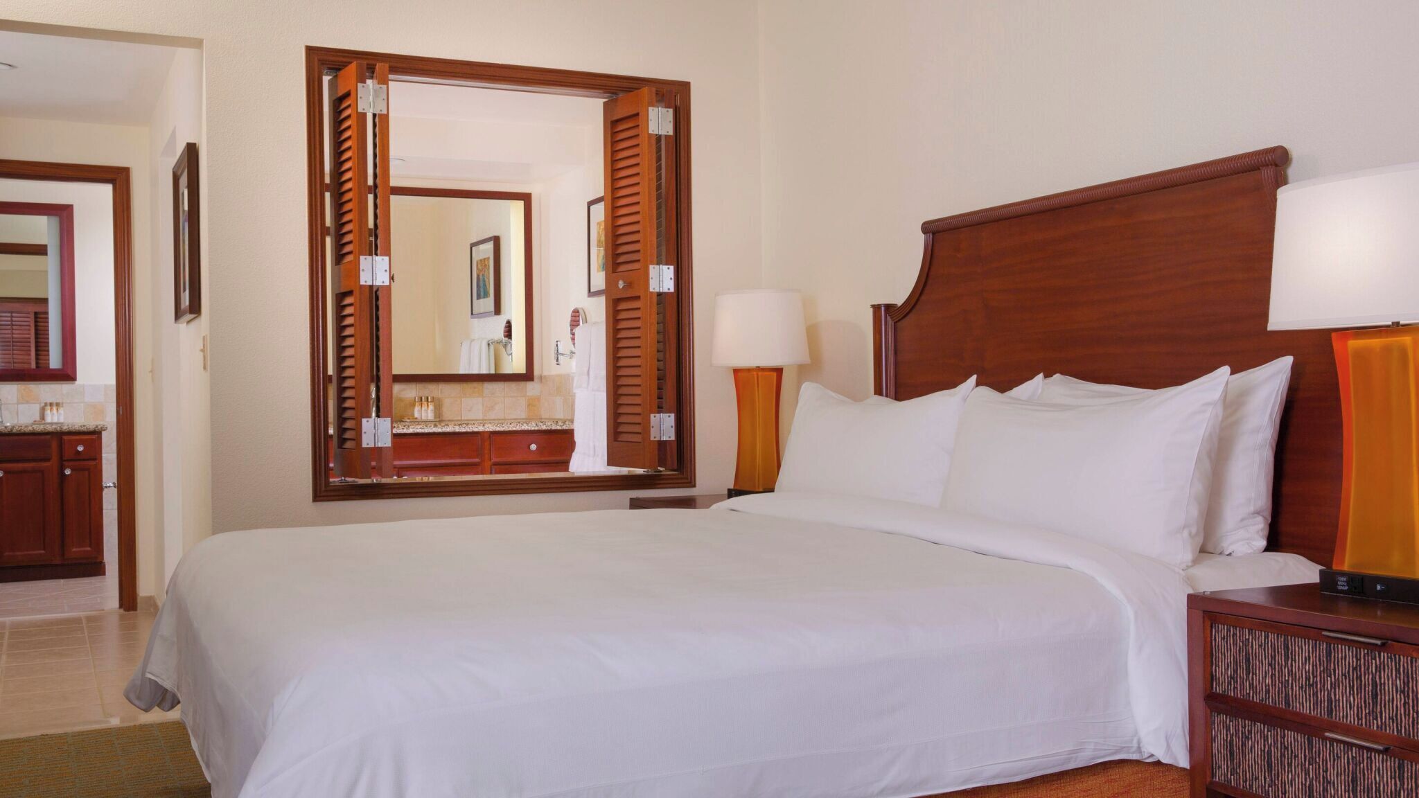 A hotel room with a large bed, a wooden headboard, a nightstand with a lamp, a mirror, and a bathroom visible through an open door.