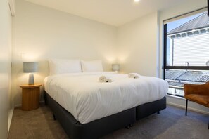 A modern bedroom with a large bed, two bedside lamps, a view of the city through a window, and a brown chair.