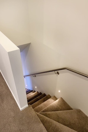 A staircase with carpeted steps and a metal handrail, leading to a landing with a white wall.