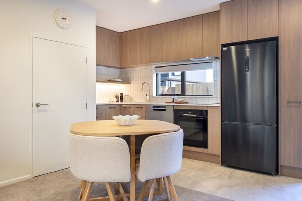 A modern kitchen with wooden cabinets, a stainless steel refrigerator, a built-in oven, and a round dining table with two chairs.