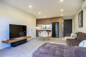 A modern living room with a flat-screen TV, a sofa, a dining table with chairs, and a kitchen area with wooden cabinets and a refrigerator.
