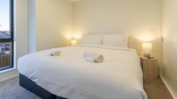 A neatly made bed with two rolled towels, a bedside lamp, and a window with a view of the outside.