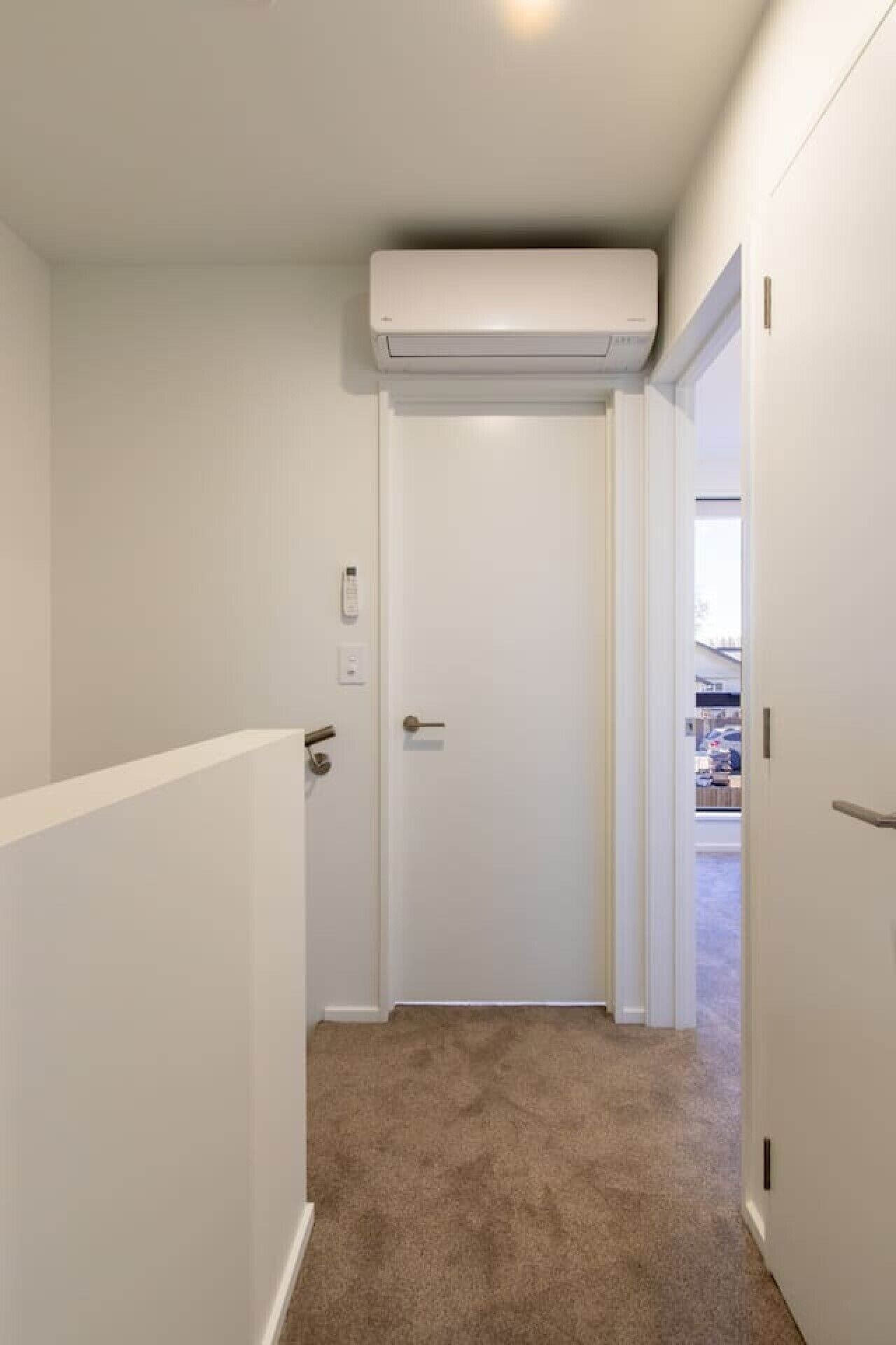 A narrow hallway with a closed door, a white air conditioning unit above, and a carpeted floor.