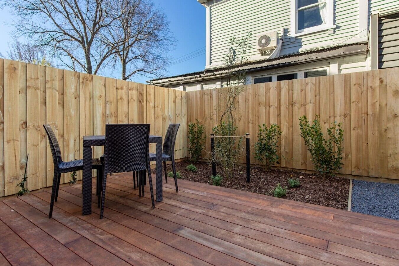 Una terraza de madera con una mesa y sillas, rodeada por un cerco de madera y plantas pequeñas.