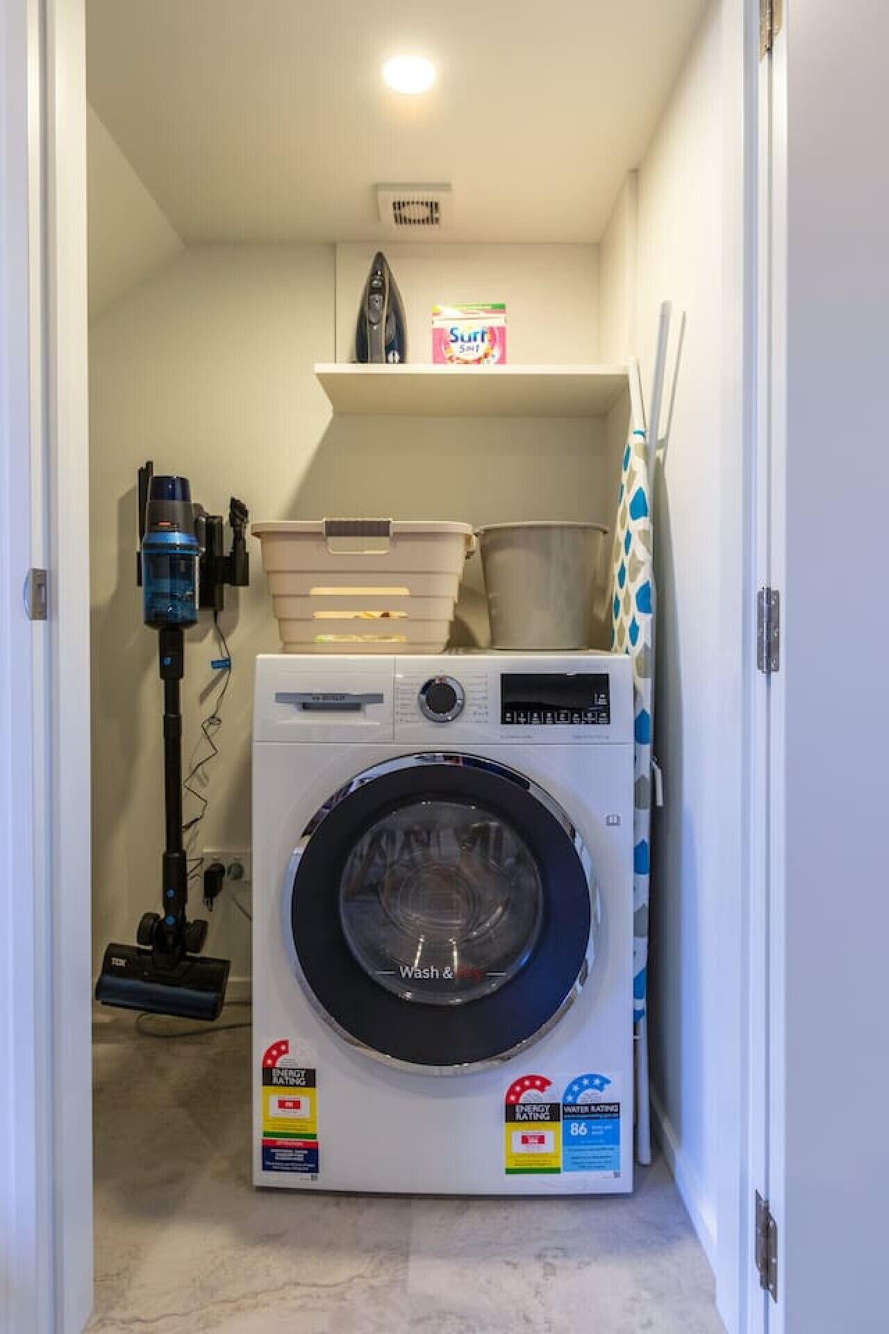 A compact laundry room with a front-loading washing machine, a vacuum cleaner, and a shelf with a box of laundry detergent.