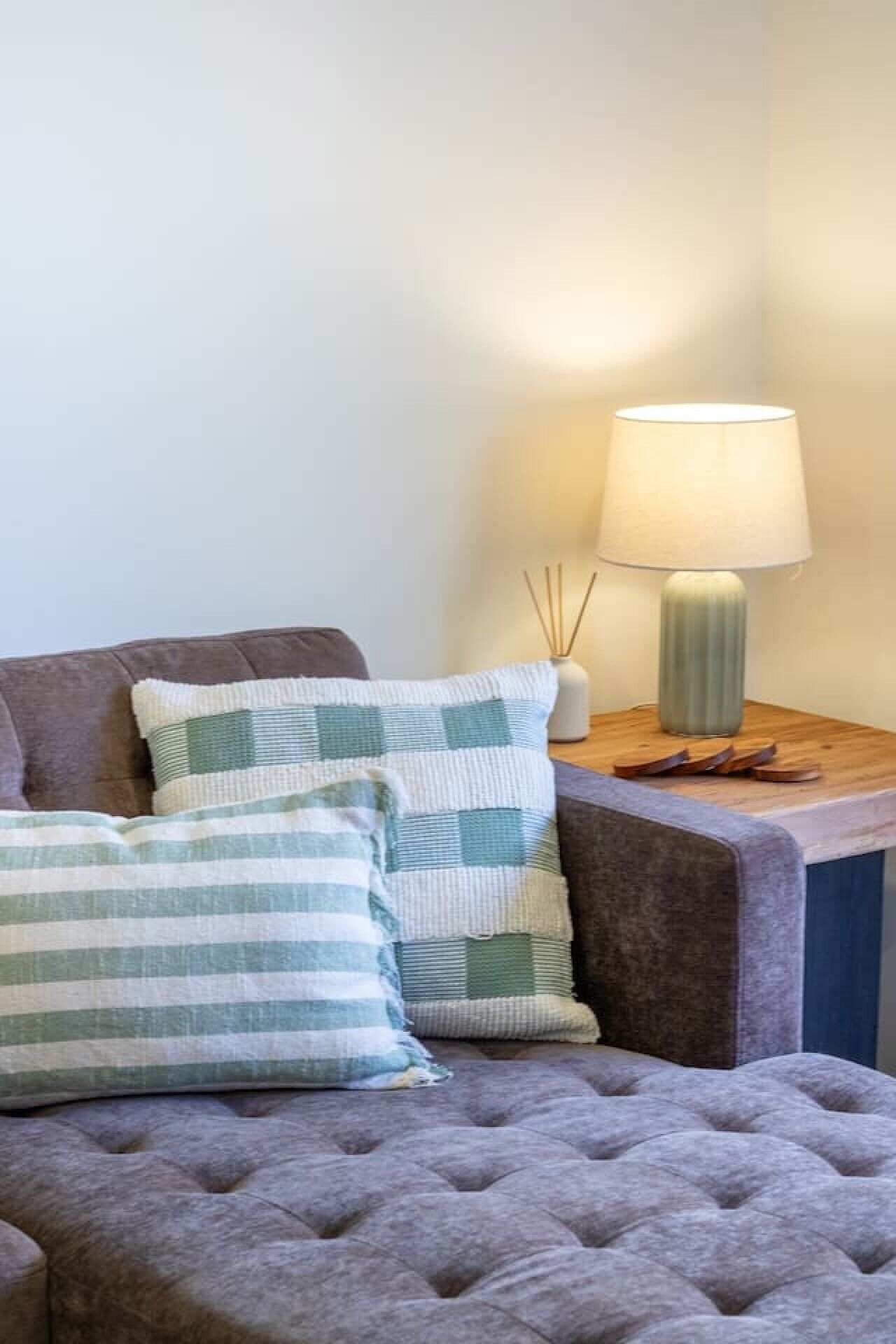 A tufted sofa with two pillows, a lamp on a side table, and a vase with reeds.