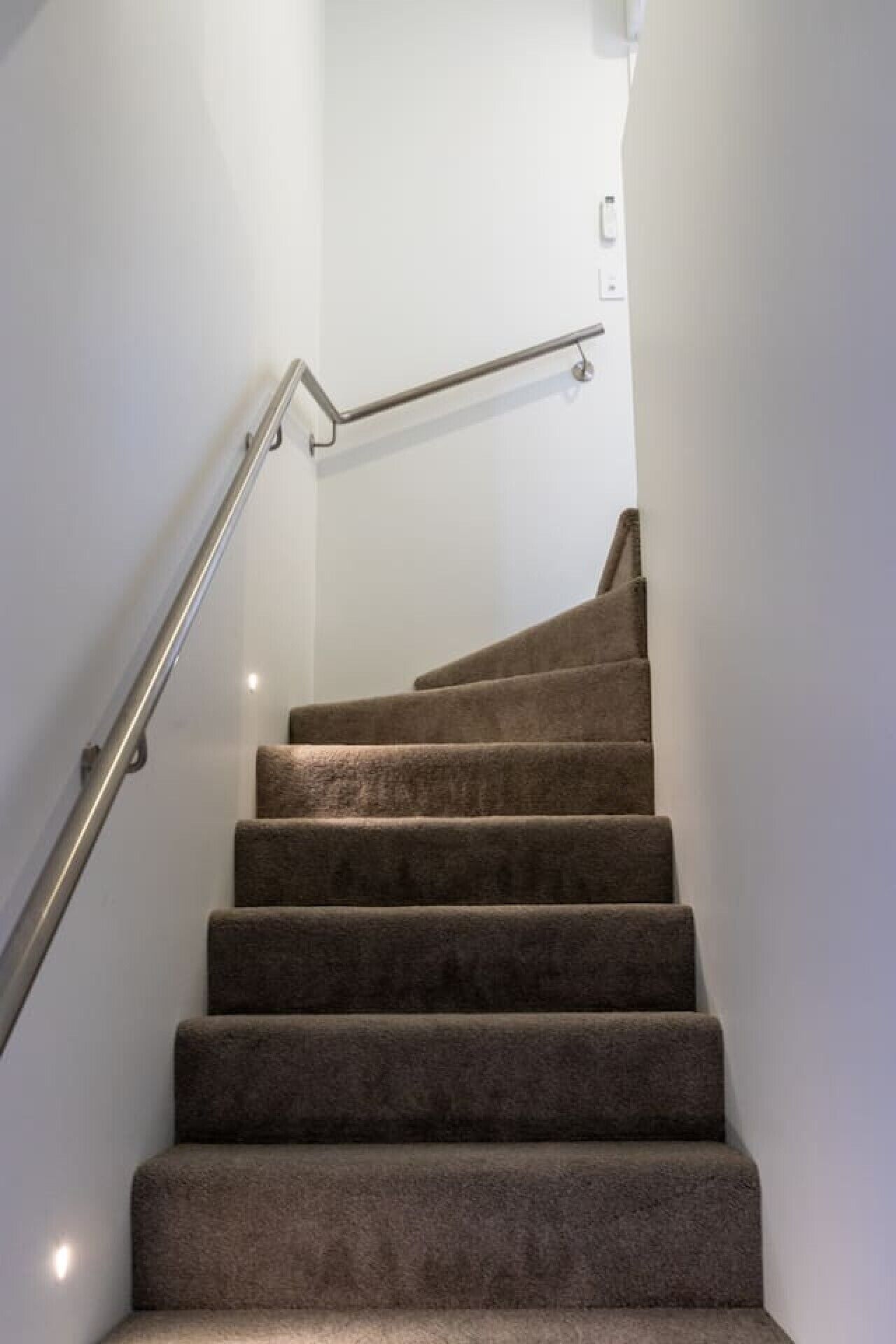 Un escalier avec des marches recouvertes de moquette et une rampe métallique, menant à un palier où se trouve un interrupteur.