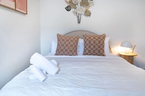 Una cama bien hecha con ropa blanca y dos almohadas estampadas, una planta en la pared y una lámpara de noche.