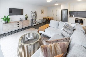 Un salon moderne comprenant un canapé d’angle, une table basse, un téléviseur et un coin cuisine avec un réfrigérateur et une cuisinière.
