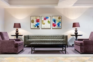 Ein modernes Wohnzimmer mit einem grauen Sofa, zwei violetten Sesseln und einem Couchtisch. An den Wänden sind abstrakte Gemälde und auf beiden Seiten des Sofas jeweils eine Stehlampe.